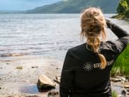 Kvinde i sort jakke med flettet hår ser ud over Loch Lomond fra stenet kyst. Grønne bakker synlige over vandet på West Highland Way.
