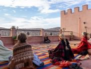 Gruppe mennesker sidder på farverige tæpper under yogasession på lyserød terrasse med udsigt til Atlasbjergene i Marokko.