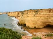 Lagdelte gyldne sandstensklipper med naturlig huleåbning langs Algarvekysten. Grøn vegetation på klippe, sandstrand nedenfor.