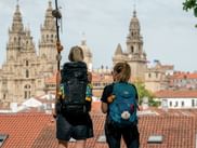 To vandrere med rygsække ser Santiago de Compostela-katedralen fra et tag. Katedralens tårne rejser sig over røde tegltage.