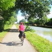 Cyklist i lyserød trøje kører langs en træbevokset kanalsti med grønt vand og frodig vegetation på begge sider.
