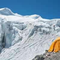 Bright orange mountaineering tent on rocky ground with massive white glacier and ice formations of Mera Peak rising behind under clear blue sky.