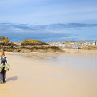 Person med ryggsäck går på bred sandstrand i Cornwall med vita kustbyggnader på klippor och stad i bakgrunden under blå himmel.