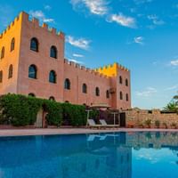Traditionel marokkansk kasbah med tinder og buede vinduer ved siden af en blå pool. Bygningen reflekteres i vandet under klar himmel.