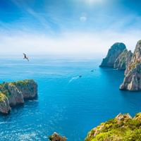 Dramatic coastal view of Capri's famous Faraglioni rock formations rising from turquoise Mediterranean waters, with green vegetation on cliffs.