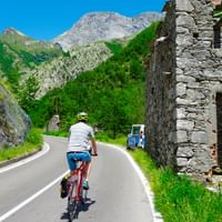 Cyklist med hjelm kører på asfalteret bjergvej forbi stenruiner, med grønne bjerge og blå himmel i baggrunden.