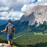 Vandrer med rygsæk og vandrestave ser på dramatiske Zugspitze toppe, grønne dale og skove under overskyet himmel.