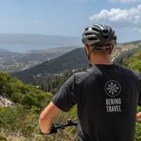 Cyklist med hjelm og sort trøje står på bakke og ser ud over grønne dale og bjerge på Kefalonia, Grækenland under skyet himmel.