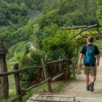 Fotturist med blå ryggsekk på tre-fjellsti med rekkverk. Frodig grønn skog dekker åssidene i Dolomittene.