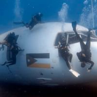 Flere dykkere udforsker et stort hvidt sunket fly under vandet i Aqaba, Jordan. Flyet hviler på den sandede havbund med klart blåt vand.