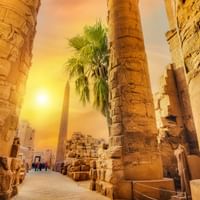 Massive steinsøyler med hieroglyfiske utskjæringer ved Karnak-templet i Egypt, opplyst av gyllent solnedgangslys med palmer.