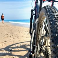 Nærbilde av terrengsykkelhjul med pigger på sandstrand i Albania. Person går i det fjerne ved turkist hav og klipper.