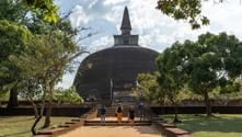 Fyra besökare går mot en stor gammal buddhistisk stupa i Polonnaruwa, Sri Lanka, inramad av gröna träd under en molnig himmel.