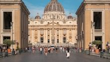 St. Peter's Basilica in Rome with its iconic dome framed by symmetrical buildings. Tourists walk along Via della Conciliazione under blue sky.