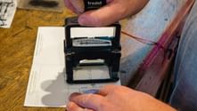 Hænder der bruger et Trodat-stempel på en pilgrimslegitimation til Camino de Santiago, med papirer og træoverflade synlige.