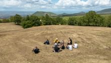 Group of hikers resting in a circle on a dry field along the Via Francigena trail. Rolling hills and green valleys visible under cloudy sky.