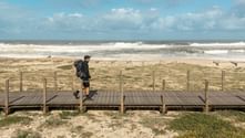 Hiker with backpack walking on wooden boardwalk along Portuguese Camino coast. Sandy beach with waves and wooden fences visible in background.