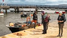 Group of hikers with backpacks at a dock preparing to board a small boat. A bridge spans the water in the background under cloudy skies.