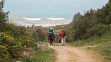 Three hikers with backpacks walking on a dirt path through green vegetation on the Portuguese Camino, with cloudy sky and distant hills.