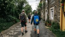 Tre pilgrimme med rygsække går på en stenbro forbi traditionelle huse. En bærer en blå jakke med Camino-muslingesymbol.