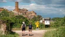 Two hikers with backpacks walking on a dirt path toward the hilltop town of Orvieto with its distinctive bell tower and historic buildings.