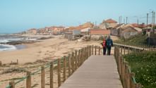 Two hikers with backpacks walking on wooden boardwalk along Portuguese coast. Sandy beach and coastal village with orange roofs visible.
