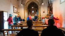 Interiør af stenkirke med pilgrimme med rygsække siddende i træbænke. Udsmykket alter med religiøse malerier synligt forrest.