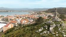 Panoramic view of Portuguese coastal town with orange-roofed buildings, harbor, and bay. Stone cross monument on rocky hill in foreground.