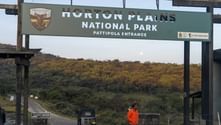 Ingångsskylt för Horton Plains nationalpark Pattipola-ingång på Sri Lanka, med skogklädda kullar i bakgrunden och en person i orange jacka.