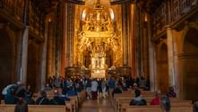 Interior of Santiago de Compostela Cathedral with pilgrims seated in wooden pews facing the ornate golden Baroque altarpiece.