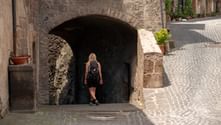 Female hiker with backpack walking through ancient stone archway on cobblestone street along Via Francigena between Orvieto and Rome.