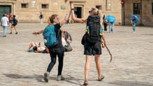 Two female hikers with backpacks high-fiving on a stone plaza. Historic building in background with other pilgrims resting and walking.