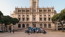 Large blue Porto sign in plaza with tourists in front of ornate beige historic building with clock tower, flanked by green trees under clear sky.