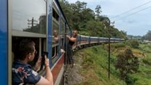 Passagerare lutar sig ut från öppna tågdörrar på ett blått och vitt tåg genom gröna kullar i Sri Lanka, med frodig vegetation synlig.