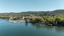 Aerial view of an Italian lakeside town with a marina, red-roofed buildings, and green hills. Clear blue sky above the scenic landscape.