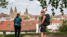 Two hikers with backpacks on a hill overlooking Santiago de Compostela. The cathedral towers dominate the skyline above red-tiled roofs.