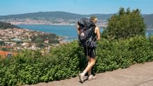 Hiker with backpack and walking stick on Portuguese Camino, overlooking bay with coastal town and green hills under blue sky.