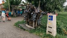 Stenkilometersten med gult Camino-muslingesymbol ved træ dækket af metallåse. Pilgrimme med rygsække hviler ved nærliggende café.