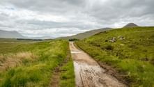 Våd grusvej på West Highland Way gennem grønt hedelandskab med bakker under overskyet himmel i Skotland.