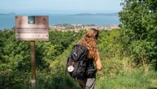 Female pilgrim with backpack standing by Camino de Santiago sign, overlooking coastal town and blue sea with islands in Portugal.