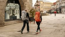 Two hikers with backpacks walk through a stone-arched plaza in Portugal. Historic buildings and a fountain are visible in the background.