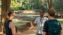 Guide med blått nyckelband talar med två turister vid de antika ruinerna i Sigiriya, Sri Lanka, med stenstrukturer och träd synliga.