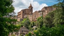 Italian hilltop town with stone buildings and church bell tower framed by green trees. Traditional architecture on the Via Francigena route.