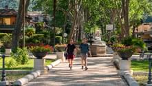 Two hikers walking on tree-lined path with flower planters in Italian town on Via Francigena route to Rome.