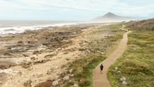 Lone pilgrim walking on coastal path beside rocky shore on Portuguese Camino. Conical mountain visible in misty distance across the ocean.