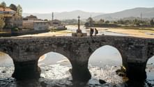 Two pilgrims walking on ancient stone bridge with three arches over shallow river. Stone cross monument stands on bridge. Portuguese Camino landscape.
