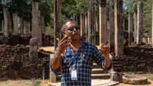 Guide med solglasögon förklarar vid arkeologiska platsen Polonnaruwa, Sri Lanka. Gamla stenpelare och ruiner synliga i bakgrunden.