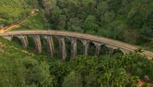 Luftfoto af en buet jernbanebro i sten med flere buer over frodig grøn tropisk skov i Sri Lanka.
