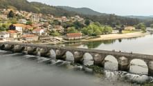 Historic multi-arch stone bridge crossing a river on the Portuguese Camino. Hillside village with red-roofed houses visible in background.