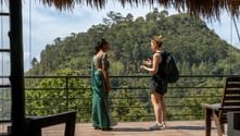 Två kvinnor på en täckt terrass med halmtak på Sri Lanka, med utsikt över grönskande kullar. En bär grön klänning, den andra shorts och ryggsäck.